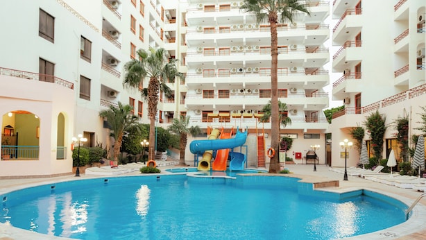 A swimming pool with a water slide, surrounded by a multi-story building with balconies and palm trees. - Empire Hotel Aqua Park (Hurghada)