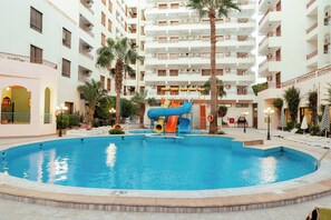 Una piscina con tobogán, rodeada por un edificio de varios pisos con balcones y palmeras.