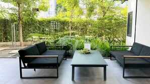An outdoor seating area with two black sofas and a central table, surrounded by lush greenery and a building in the background.