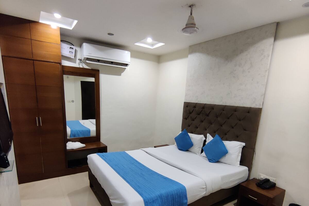 A hotel room with a bed, bedside table, nightstand, wardrobe, mirror, and a small table with a kettle, cups, and a phone.