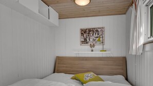 Un dormitorio con cabecera de madera, paredes blancas, un cuadro, una mesita de noche con lámpara y una ventana con cortinas.