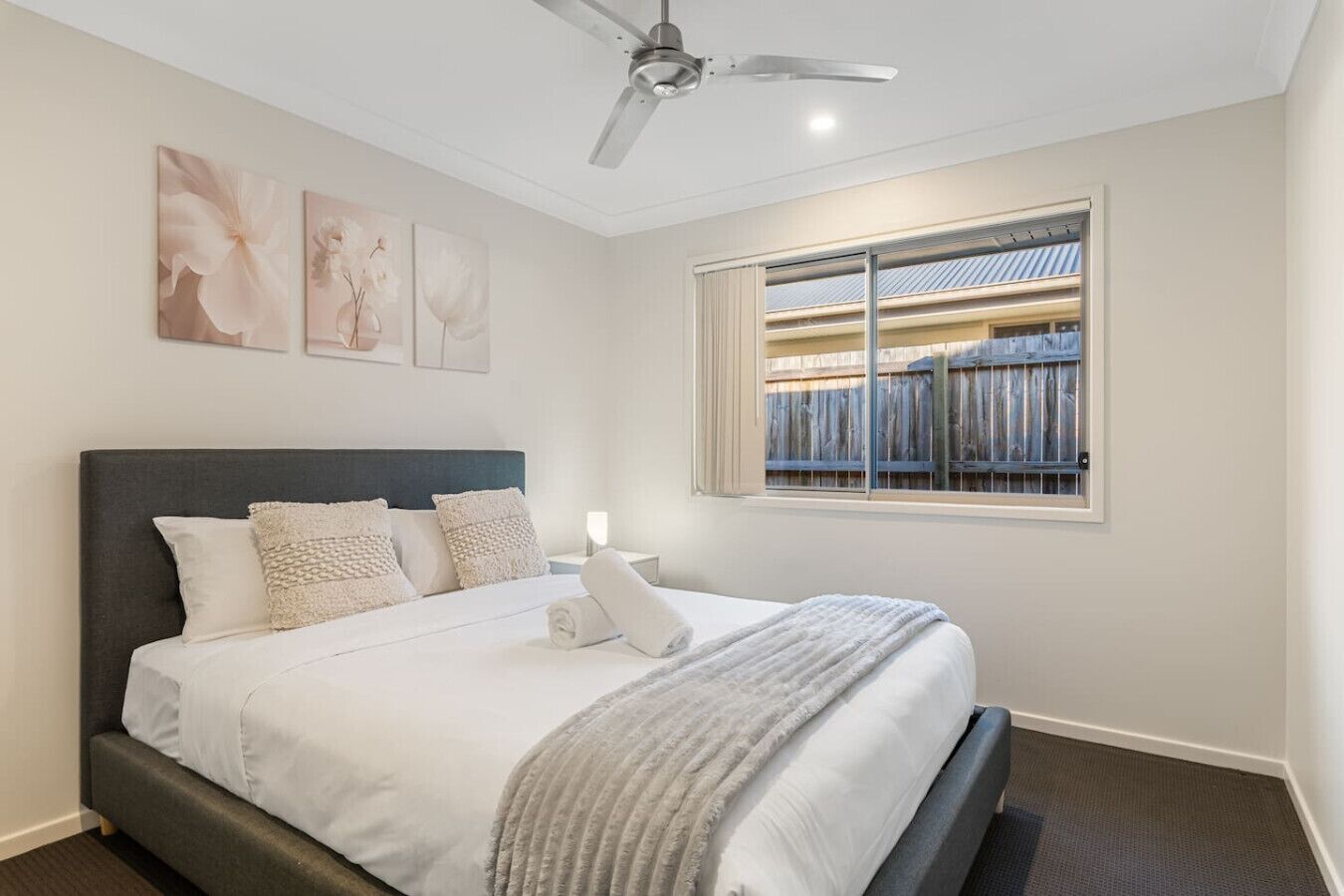 Un dormitorio ordenado con cama, almohadas, una mesita de noche y ventana con vista a una cerca de madera.