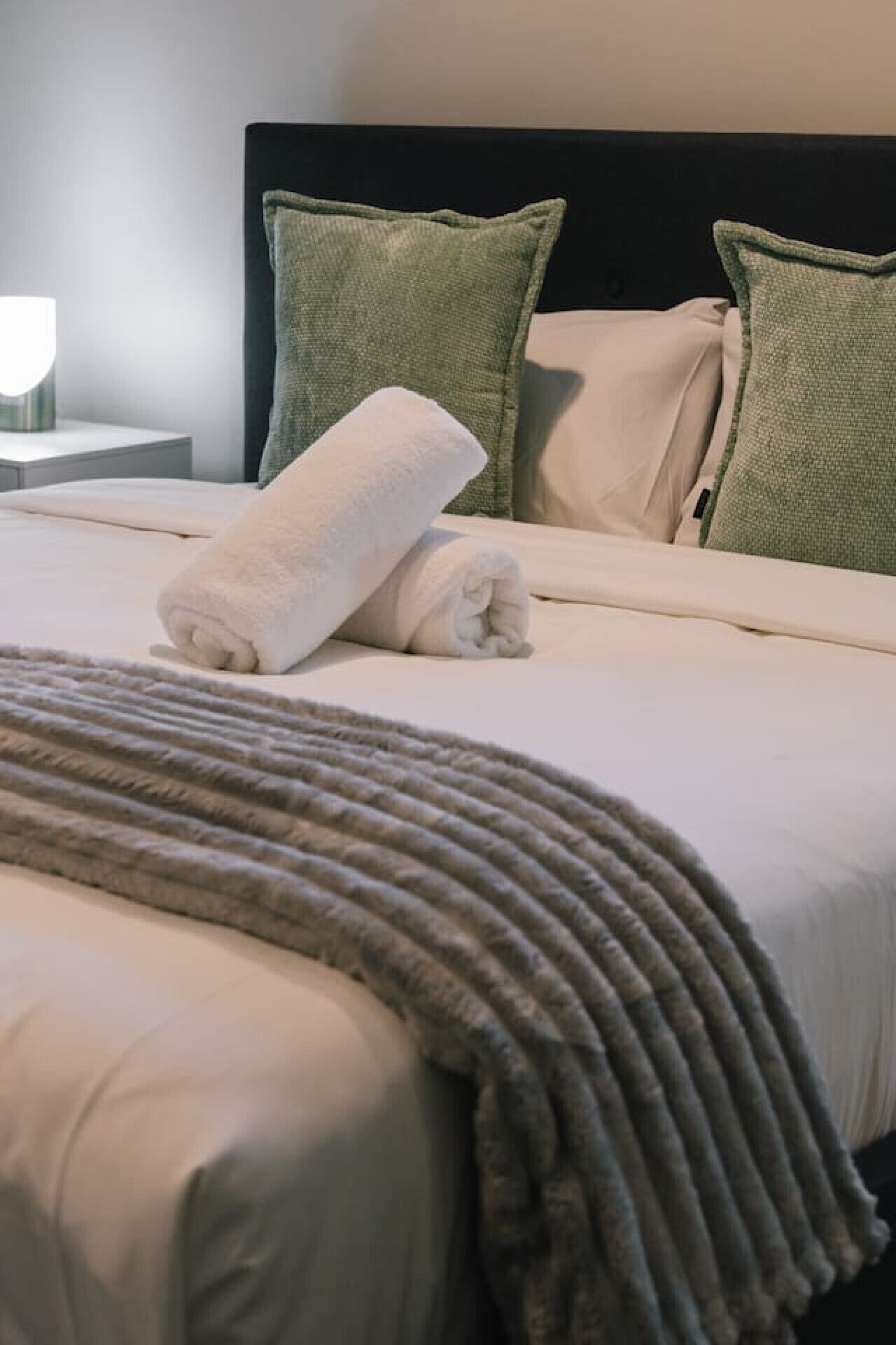 Een net opgemaakt bed met witte lakens en twee groene kussens, een grijze plaids, en een nachtkastje met een lamp.