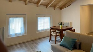 Una habitación con techo alto, vigas de madera, una mesa de comedor con sillas, un sofá y dos ventanas con cortinas translúcidas.