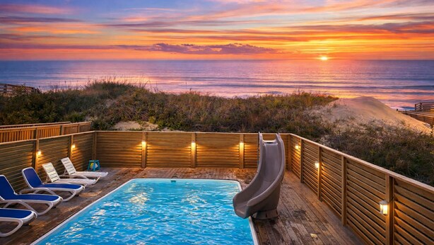 A wooden deck with a pool, lounge chairs, and a slide, overlooking the ocean at sunset.