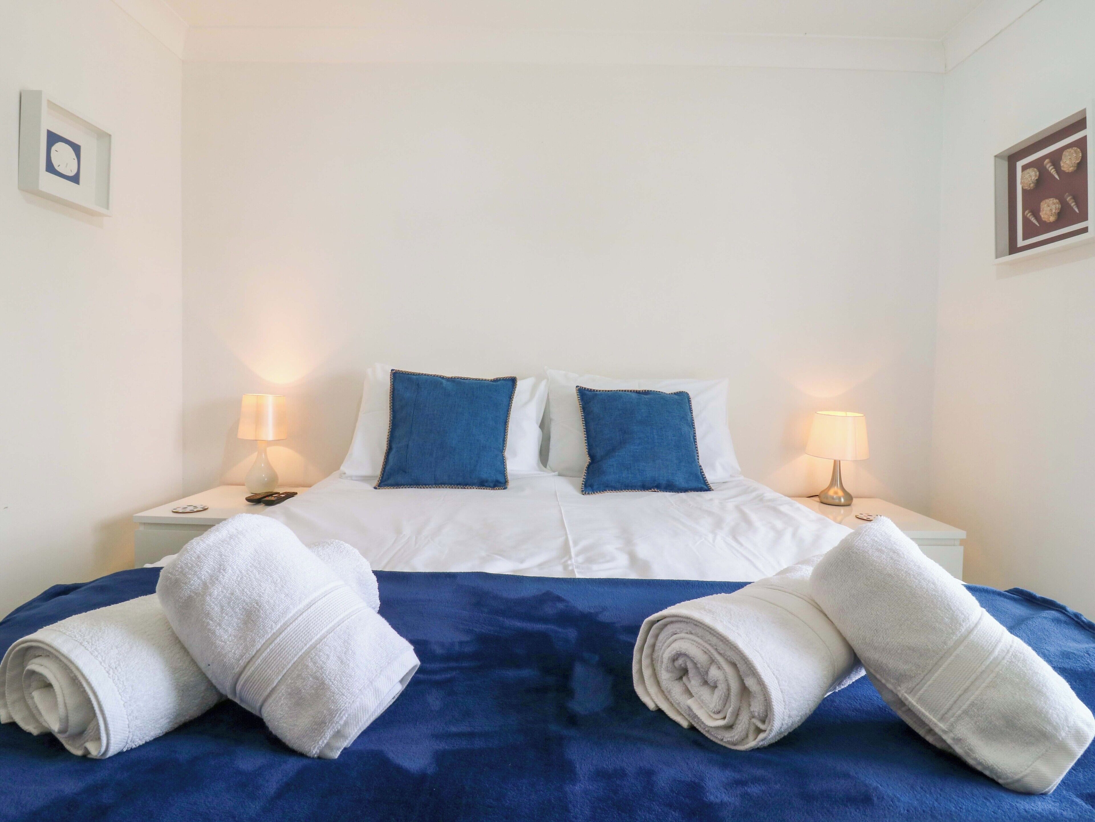 A neatly made bed with white linens and two blue pillows, two bedside tables with lamps, and a blue blanket on the bed.