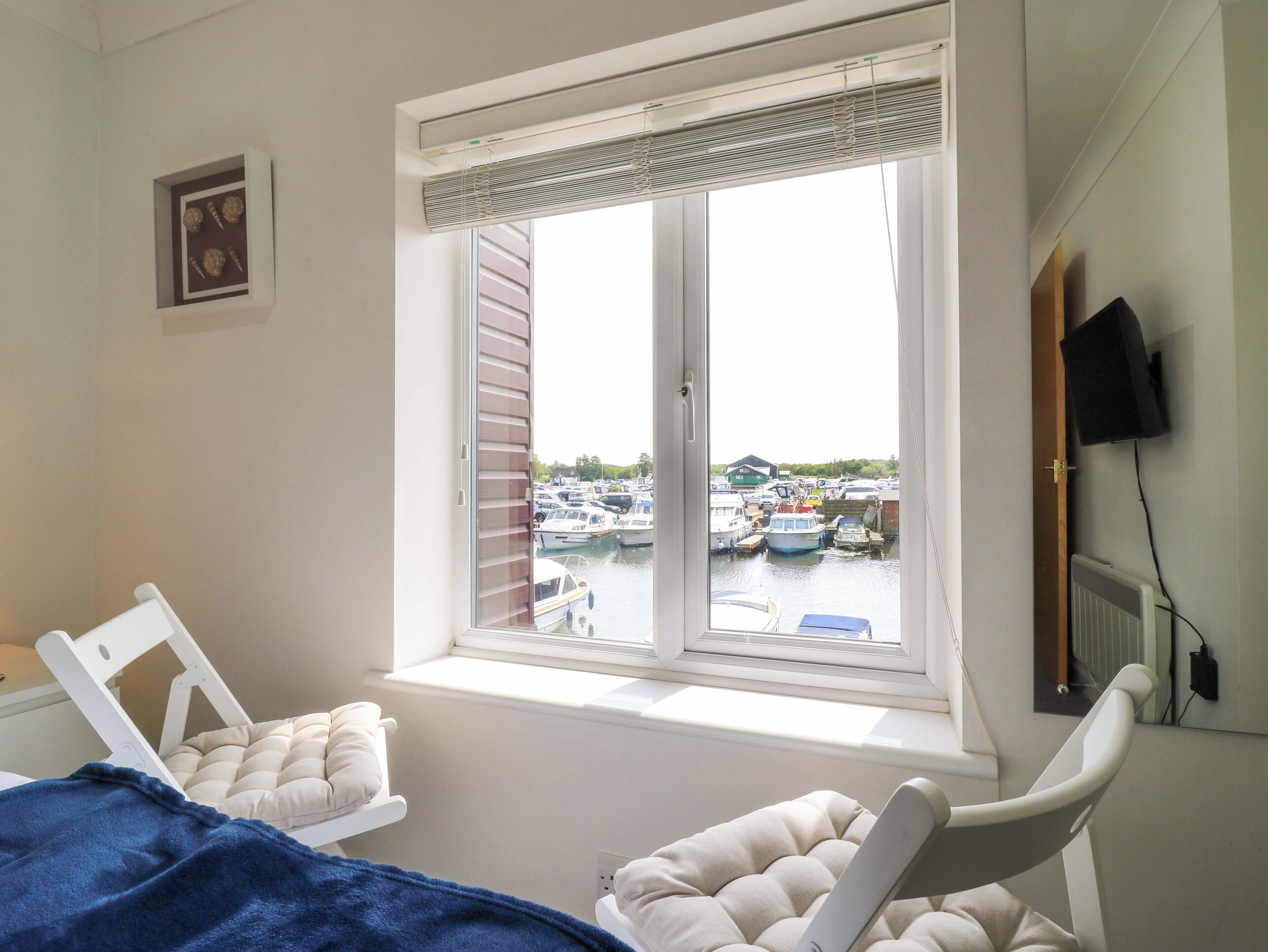A room with a large window overlooking a marina, a bed with a blue blanket, and a white chair.