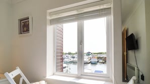 A room with a large window overlooking a marina, a bed with a blue blanket, and a white chair. - Kingsmere (Norwich)