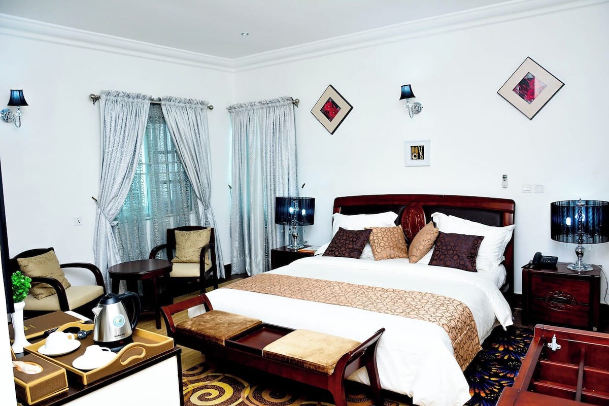A hotel room with a large bed, two bedside tables with lamps, a wooden bench, a small table with a kettle, and a chair.