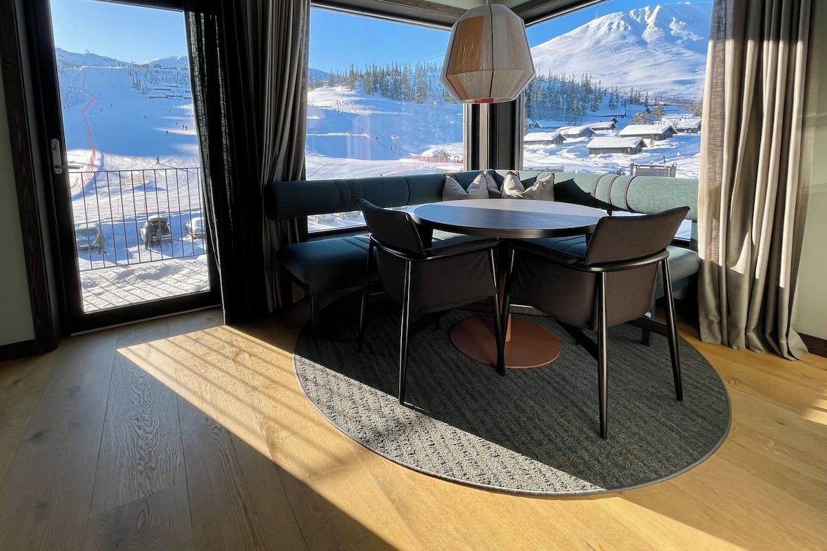 A room with a large window offering a snowy mountain view, a round table with chairs, a sofa, and a hanging light fixture.