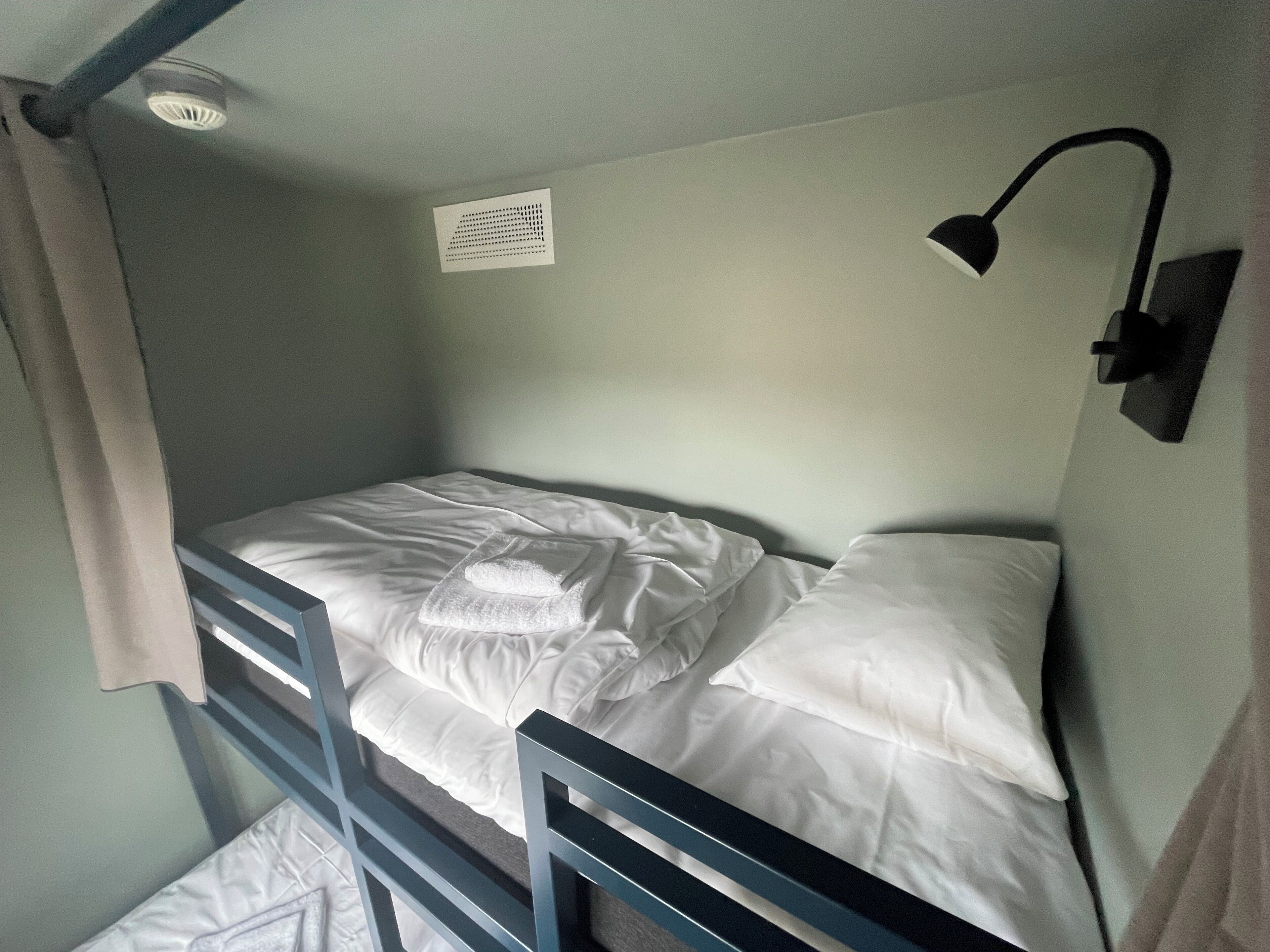 Ein Stockbett mit weißer Bettwäsche, einer Wandleuchte und einem Lüftungsgitter.