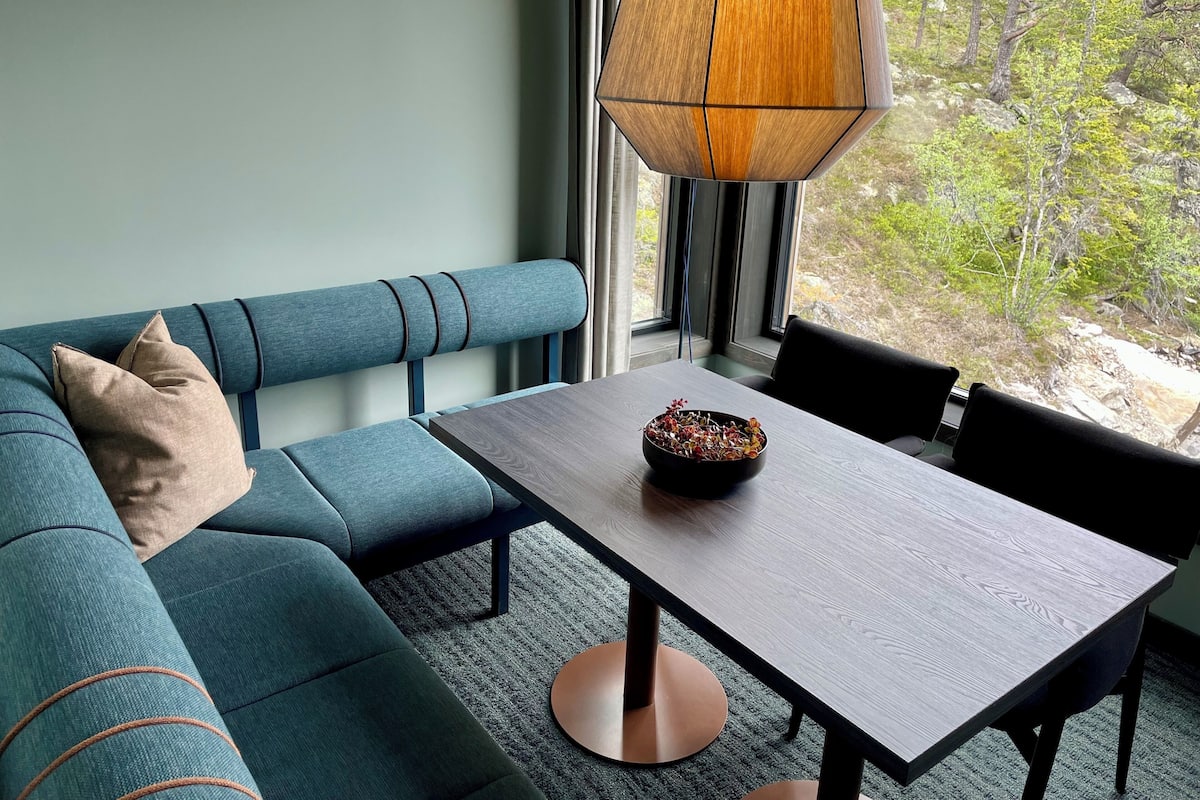 A modern living room with a teal sofa, a wooden dining table, and a view of a rocky outdoor area through a window.