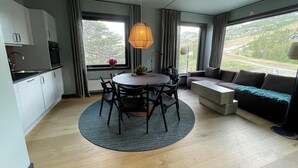 A modern kitchen with white cabinets, a dining table with chairs, a sofa, and a large window with a view of greenery. - Gausta Sportell (Tinn)