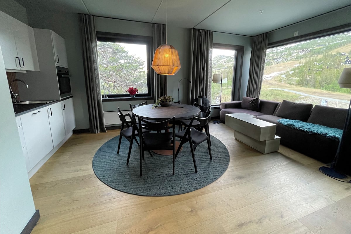 A modern kitchen with white cabinets, a dining table with chairs, a sofa, and a large window with a view of greenery.