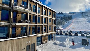 A multi-story building with balconies and large windows, located near a ski slope with parked cars and a person in ski gear. - Gausta Sportell (Tinn)