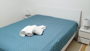 A neatly made bed with a blue bedspread and rolled white towels, a white nightstand with a lamp, and a painting of sunflowers on the wall. - Cozy vacation home in Terrasini a stone's throw from the sea and the historic center. (Terrasini)