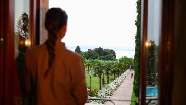 Eine Person steht an einem Fenster mit Blick auf einen Weg, viel Grün und ein entferntes Gewässer.