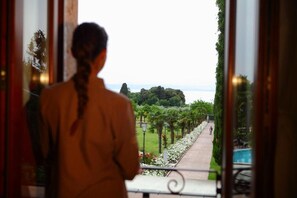 A person standing by a window overlooking a pathway with greenery and a distant body of water. - La Boheme Chambres (Lazise)