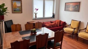 Ein Zimmer mit einem Holztisch, Stühlen, einem roten Sofa und einem gelben Sessel. Zwei Fenster mit Jalousien, ein Bild an der Wand und eine Topfpflanze links.