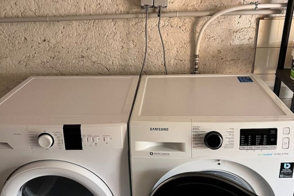 Zwei Samsung Waschmaschinen in einem Waschraum mit strukturierter Wand und freiliegenden Rohren.