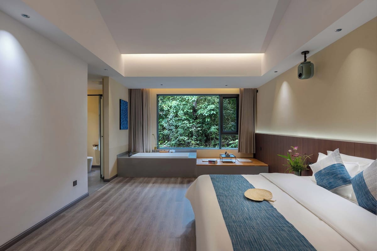 Une chambre d’hôtel moderne dotée d’un grand lit, offrant une vue sur la verdure par la fenêtre, d’un petit coin salon et d’un projecteur fixé au plafond.