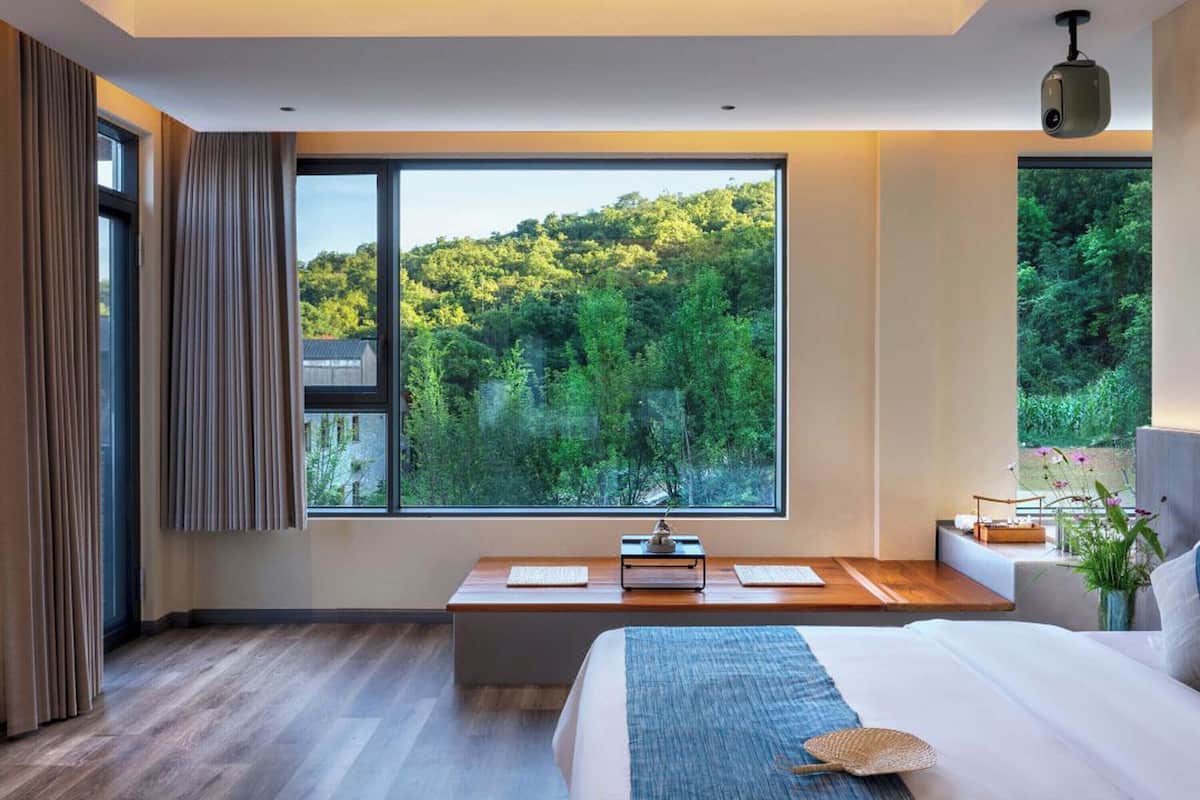 Une chambre moderne dotée d’une grande fenêtre donnant sur une forêt, d’un lit agrémenté d’un couvre-lit bleu et d’un bureau avec une lampe.