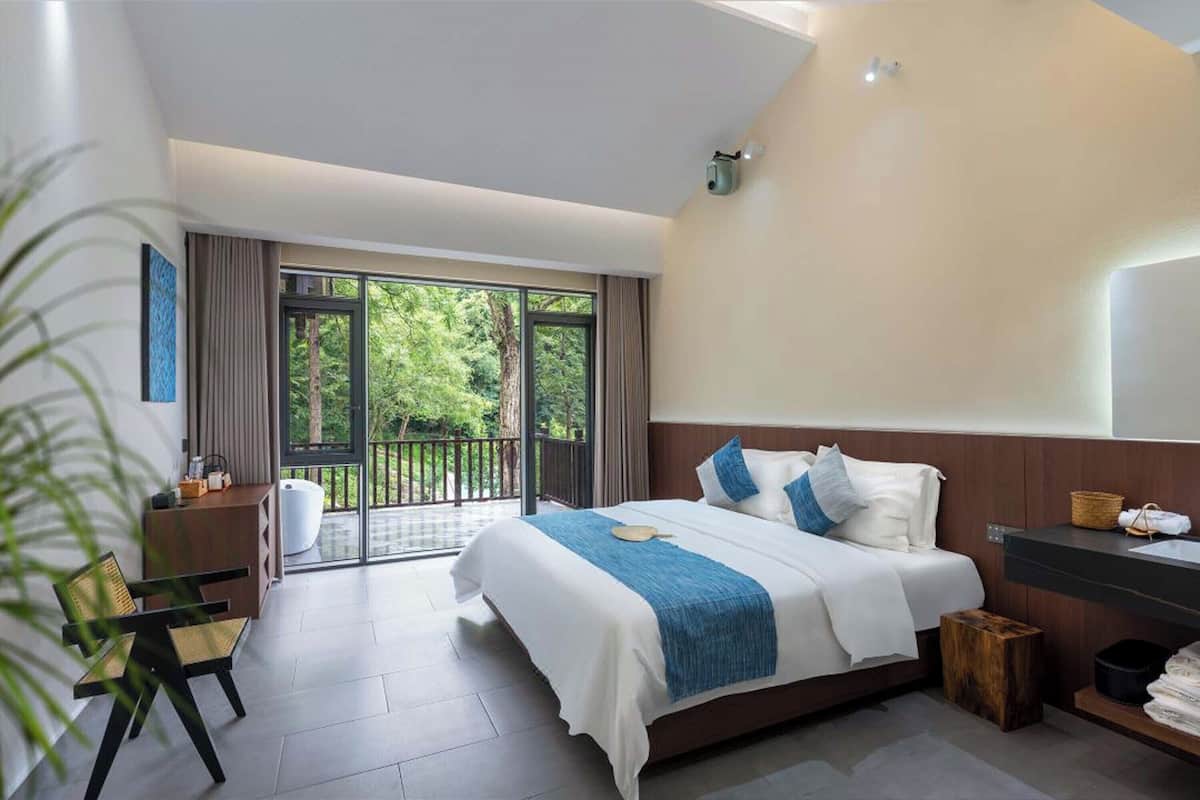 Une chambre d’hôtel moderne dotée d’un grand lit, d’une tête de lit en bois, d’un balcon avec vue sur la verdure, d’un bureau avec une chaise et d’une petite poubelle.