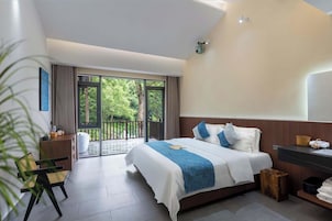 Habitación de hotel moderna con una cama grande, cabecera de madera, balcón con vistas a la vegetación, escritorio con silla y un cesto de basura peque ño.