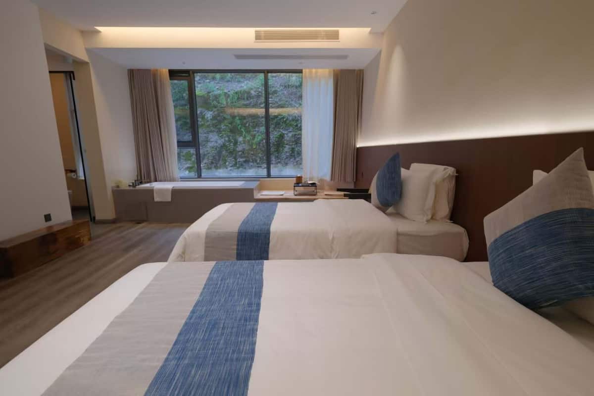 Une chambre d’hôtel avec deux lits, une fenêtre avec des rideaux, un petit coin salon et une vue sur la verdure.