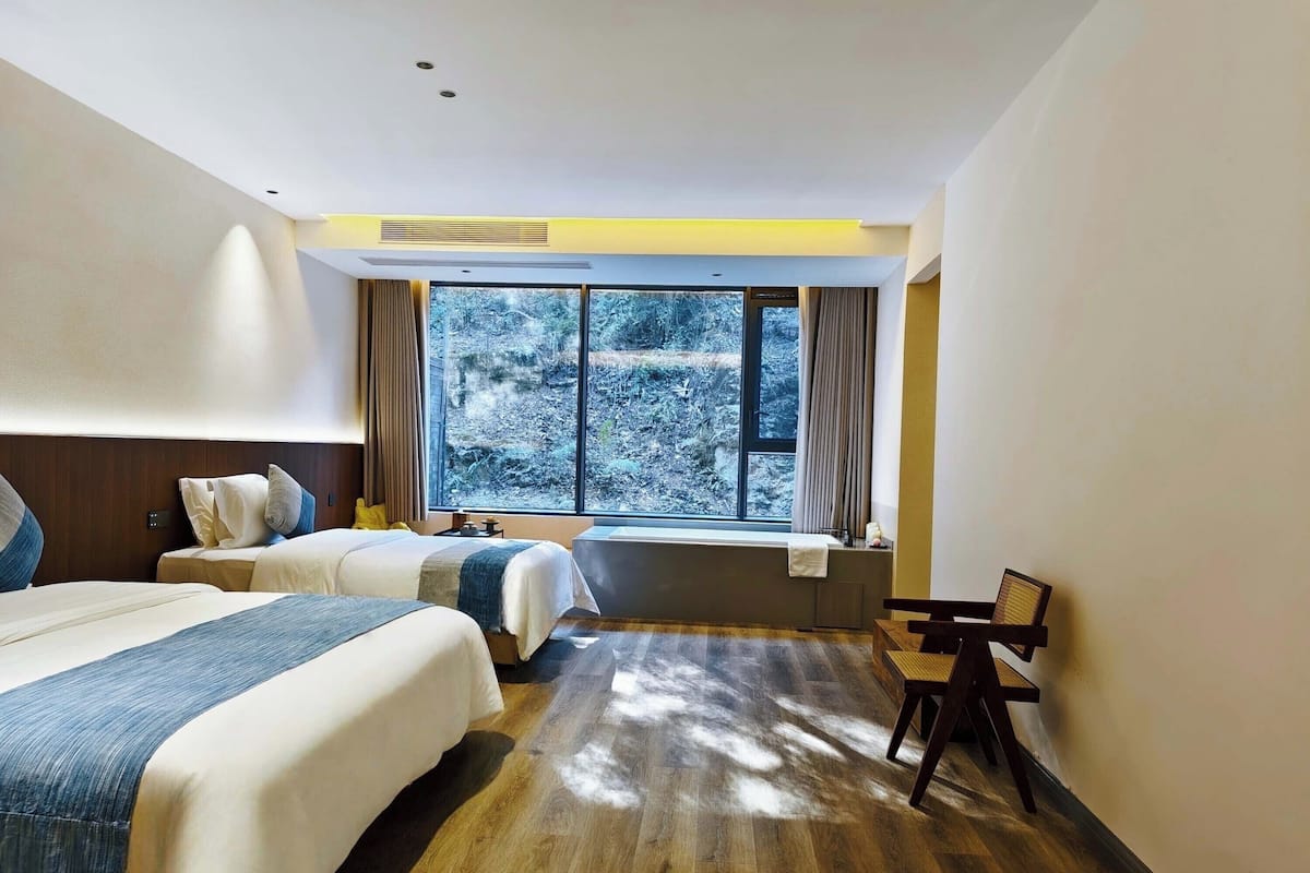 Une chambre d’hôtel avec deux lits, une grande fenêtre donnant sur la forêt, un plancher en bois et une petite table avec une chaise.
