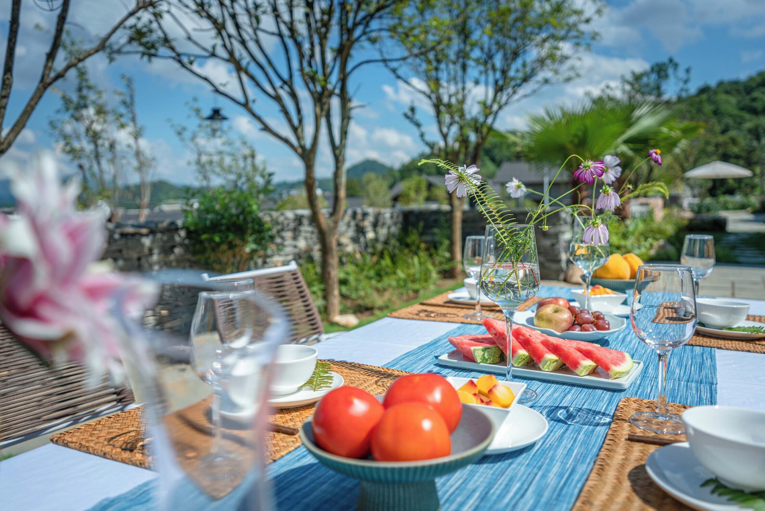 Una mesa de comedor al aire libre con mantel azul, platos de sandía y tomates, y un jarrón con flores.