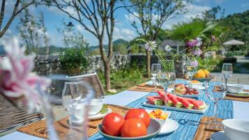 Una mesa de comedor al aire libre con mantel azul, platos de sandía y tomates, y un jarrón con flores.