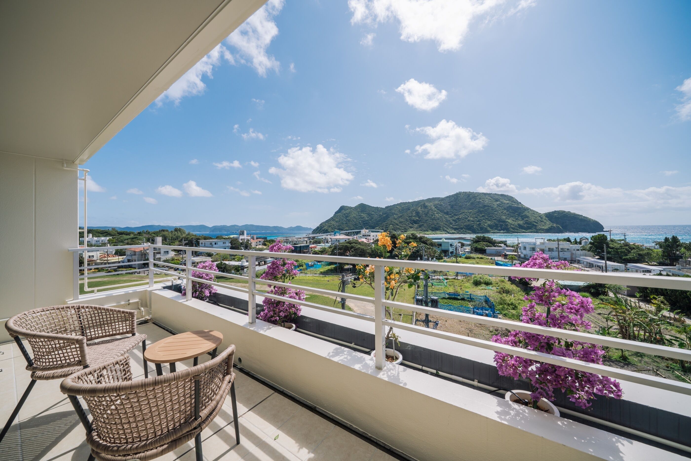 A balcony with wicker chairs and a table, overlooking a coastal area with mountains and a view of the sea.