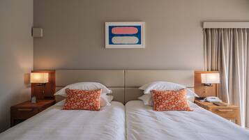A hotel room with two beds, each with white bedding and orange floral pillows. There are two bedside lamps, a framed abstract artwork on the wall, and a window with curtains.