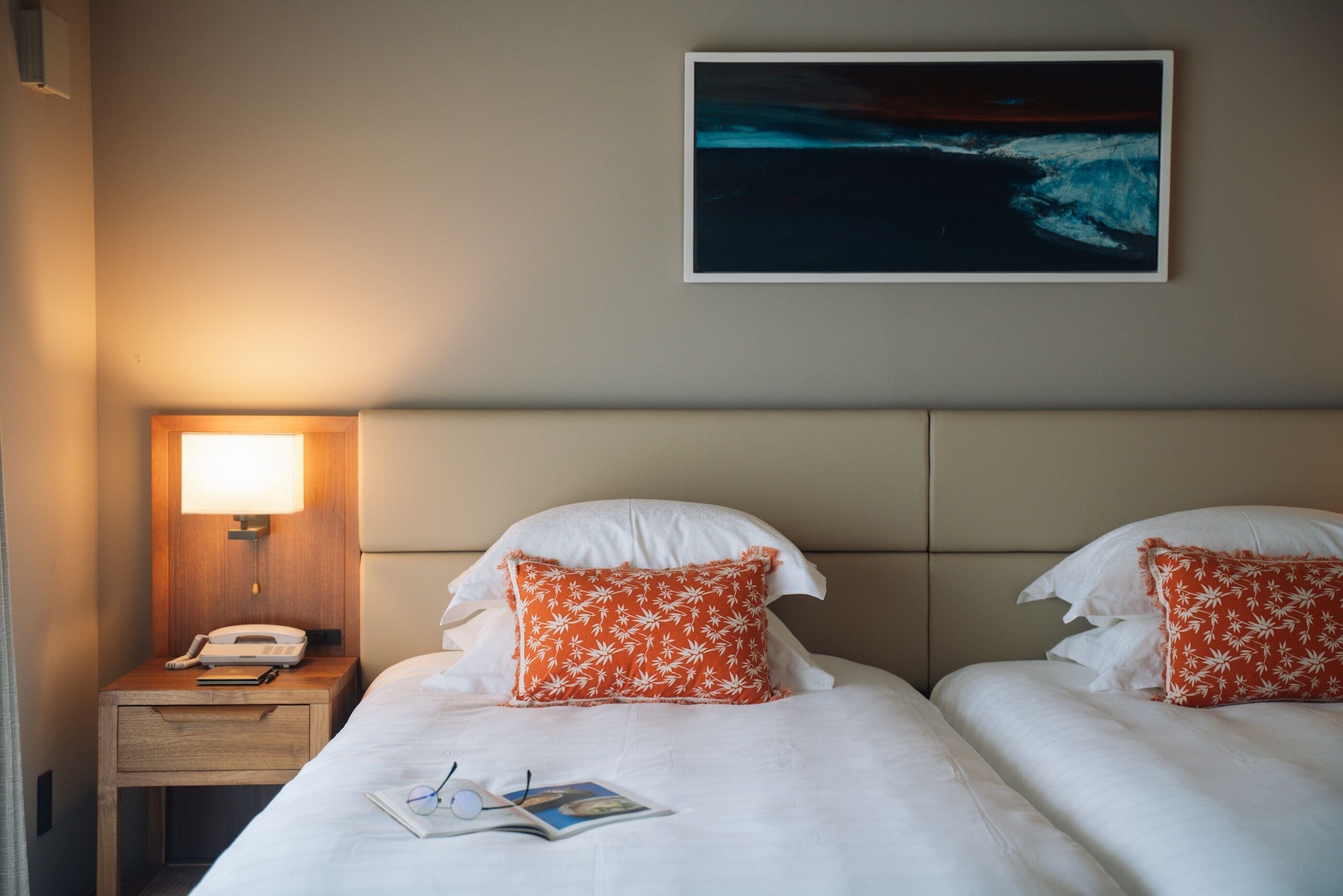 A hotel room with two beds, a nightstand, a lamp, a framed picture on the wall, and a magazine on the bed.