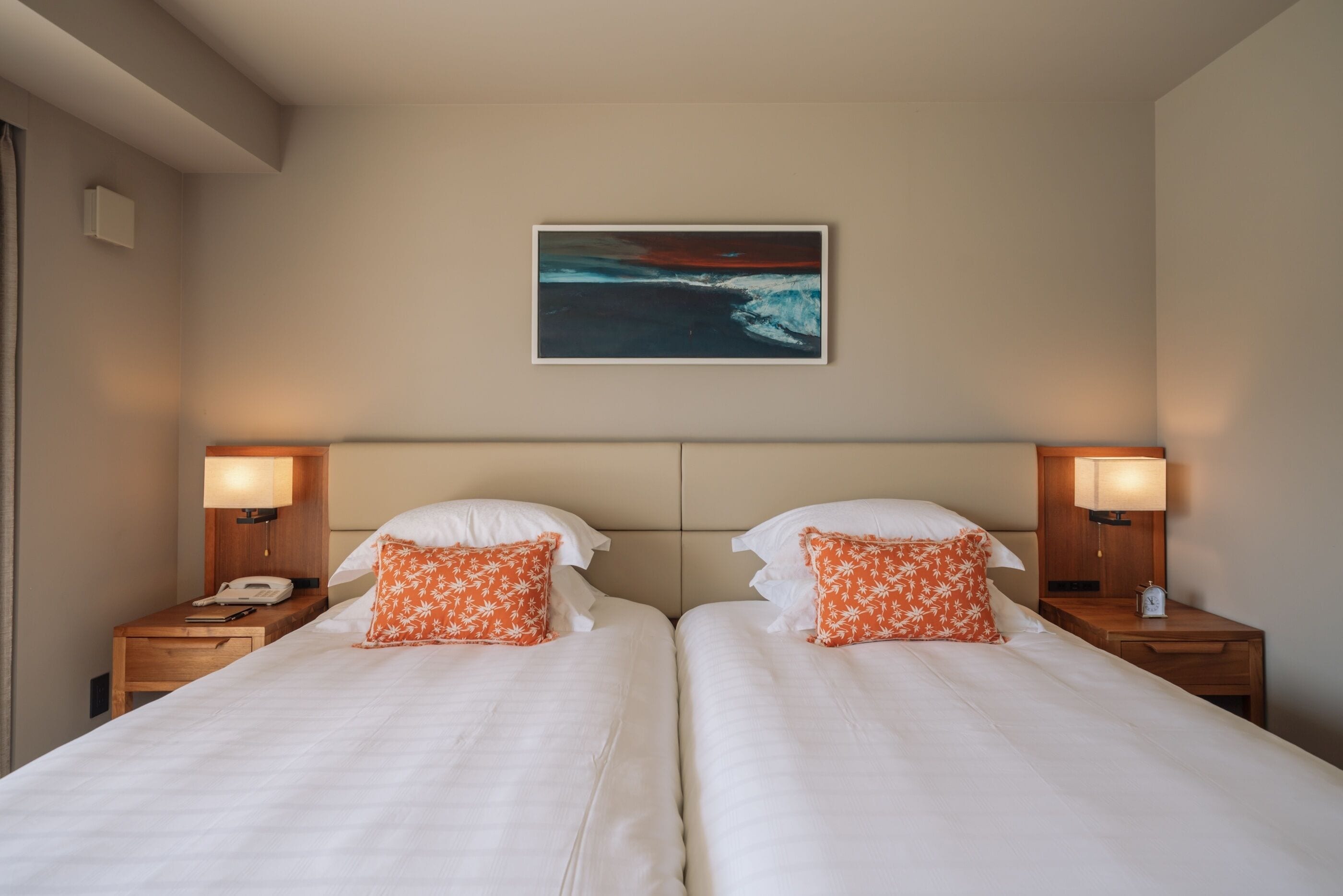 A hotel room with two beds, a painting of a seascape, bedside lamps, and a telephone on the nightstand.