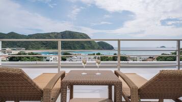 A balcony with wicker furniture and two glasses of white wine, overlooking a coastal landscape with hills and a body of water.