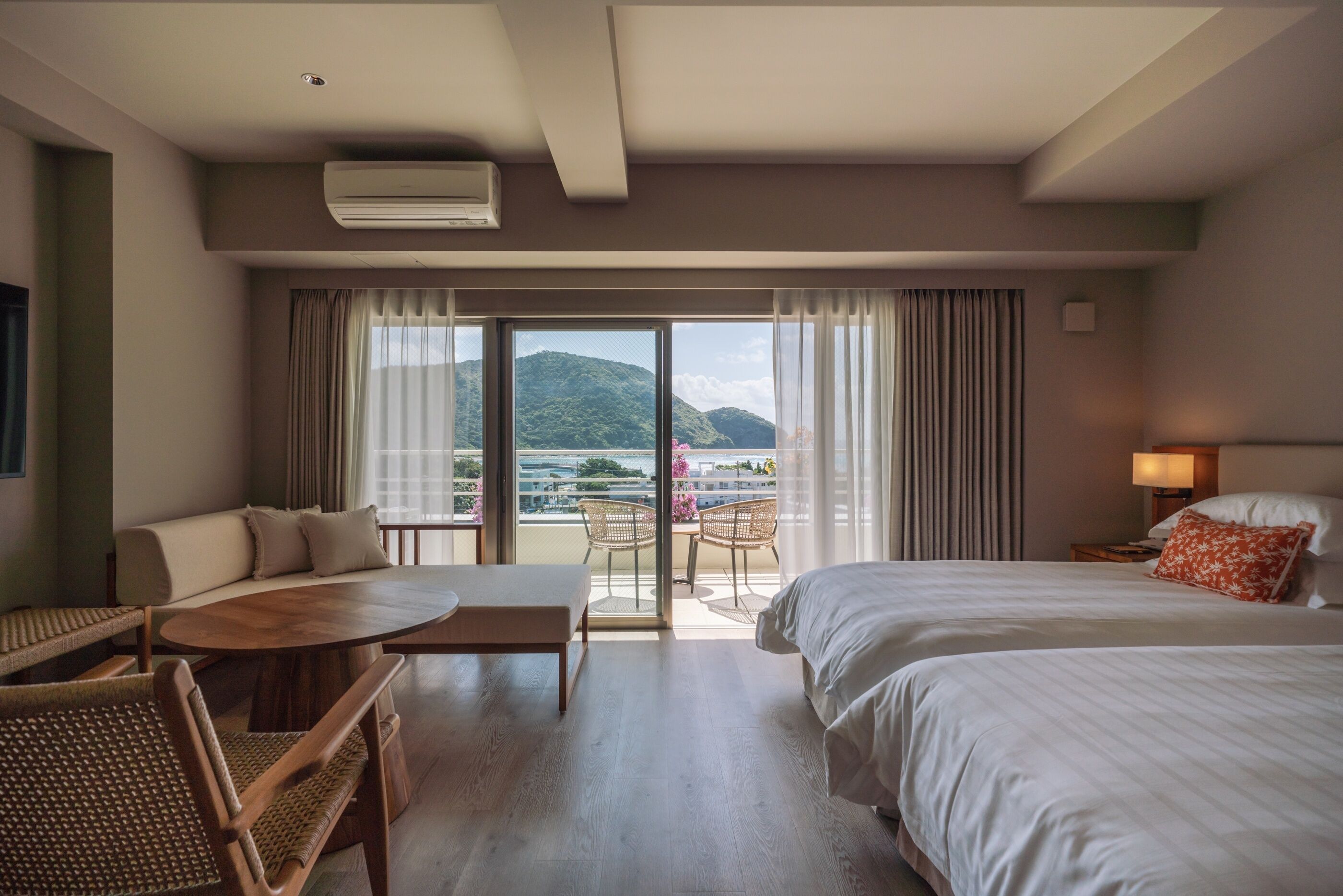 A hotel room with a bed, a sofa, a table, and a balcony with a view of the sea and hills.