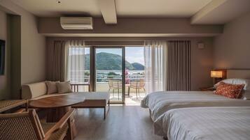 A hotel room with a bed, a sofa, a table, and a balcony with a view of the sea and hills.