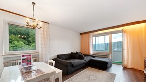 Un salon moderne avec un canapé d’angle, une table à manger et une grande fenêtre donnant sur une forêt.