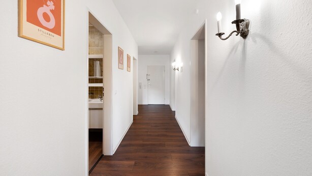 Un couloir avec un plancher en bois, des murs blancs et un panneau encadré sur le côté gauche.