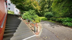 Un escalier extérieur menant à un bâtiment doté d’une rambarde en métal, entouré de verdure et bordé d’un chemin pavé.