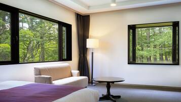 A hotel room with a bed, a sofa, a table, and large windows offering a forest view.