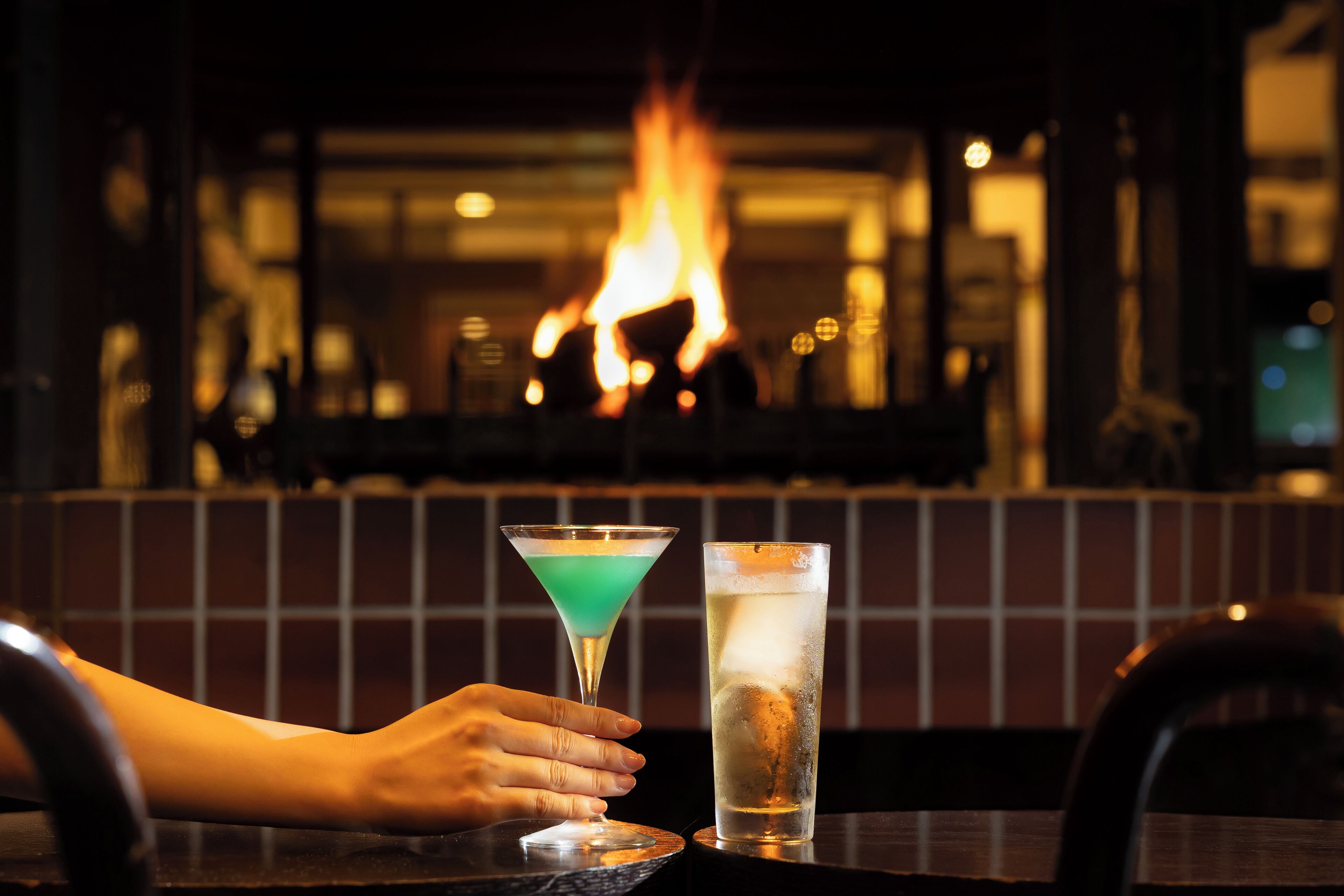 Une scène de bar avec une main tenant un verre à martini et un grand verre d’eau, et une cheminée en arrière-plan.