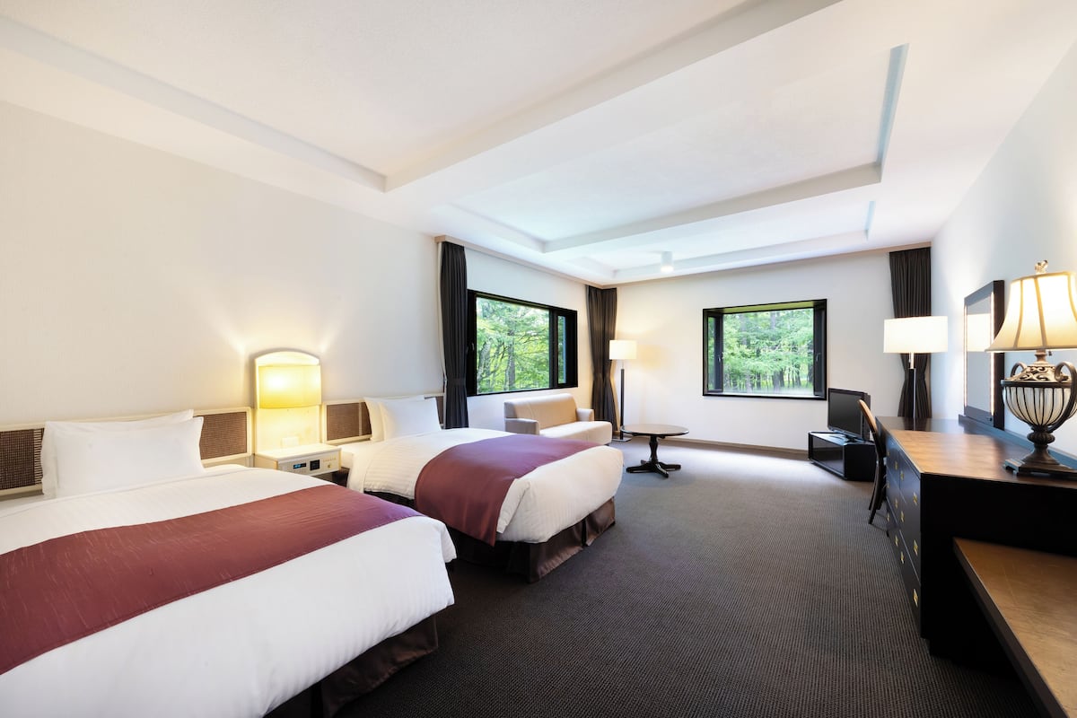 A hotel room with two beds, a desk, a chair, a lamp, and a view of greenery outside.