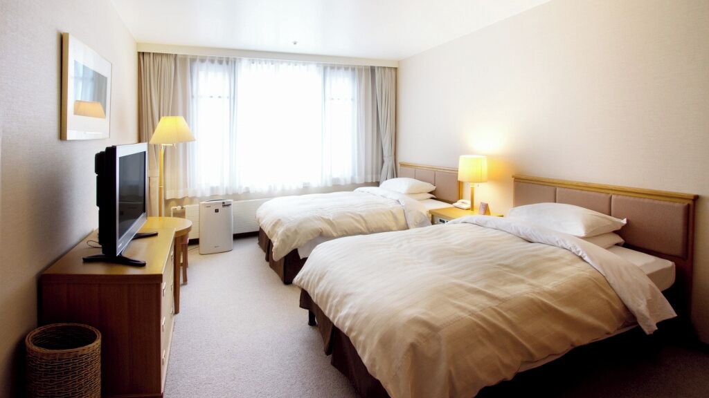Quarto de hotel com duas camas, uma mesa de trabalho, televisão, luminária e janela com cortinas.