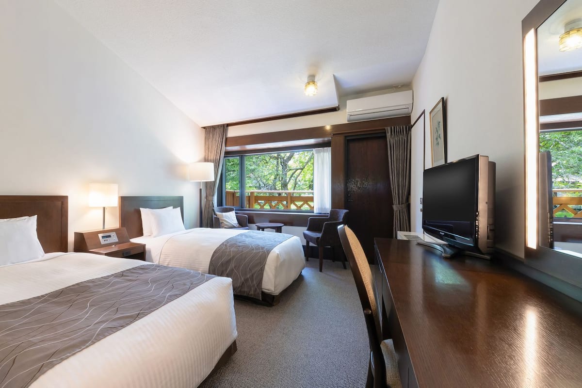Habitación de hotel con dos camas, un escritorio, un televisor y una ventana con vistas a zonas verdes.