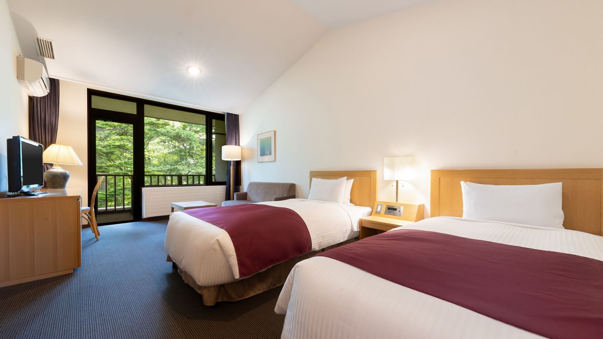 Ein Hotelzimmer mit zwei Betten, einem Schreibtisch mit Fernseher, einem Sessel, einem Fenster mit Blick auf Bäume und einem Balkon.
