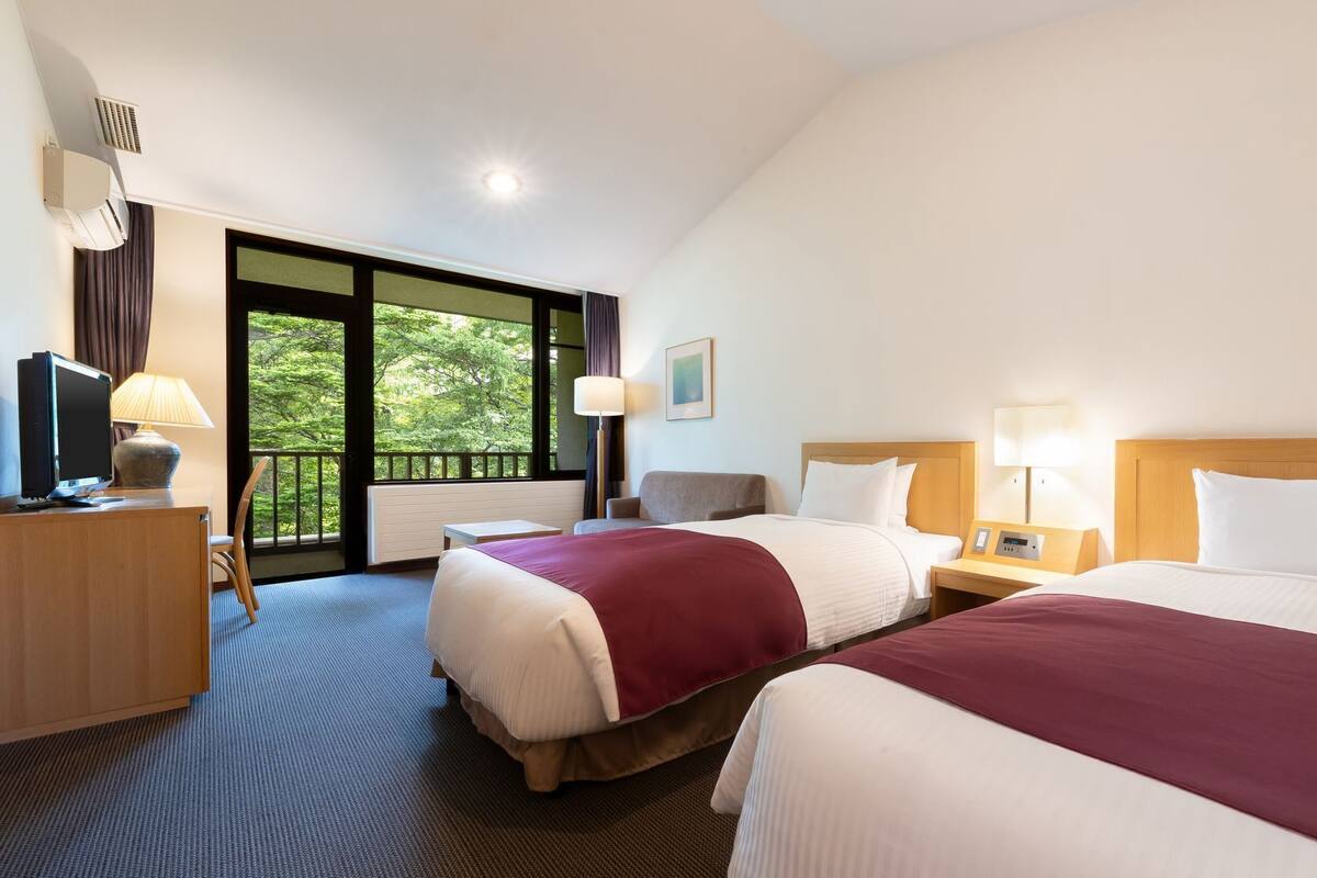 A hotel room with two beds, a desk with a TV, a chair, a window with a view of trees, and a balcony.