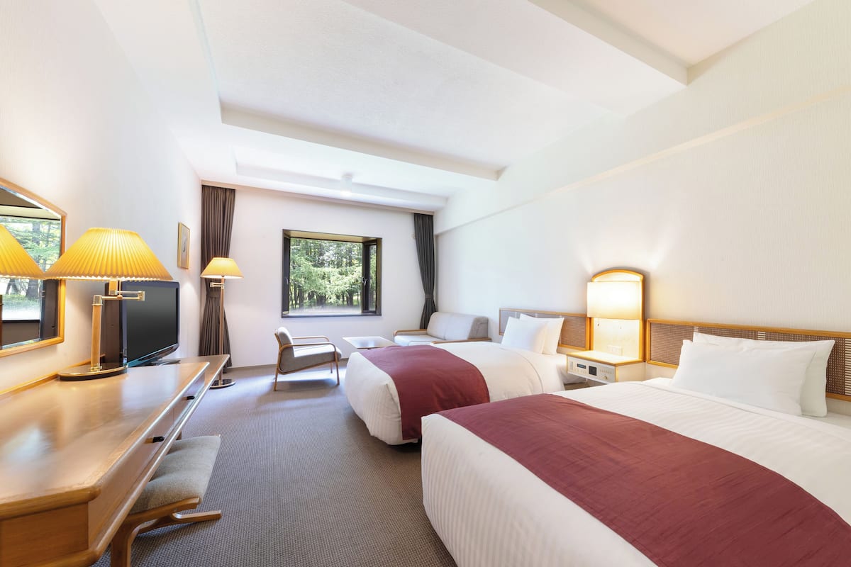 A hotel room with two beds, a desk, a chair, a TV, and a window with a view of trees.